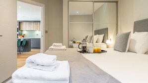 Una habitación de hotel moderna con una cama grande, una mesita de noche con lámpara, un espejo y vistas al área de la cocina.