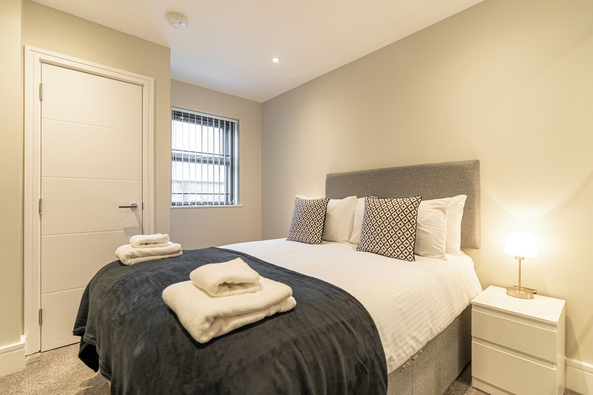 A neatly made bed with a dark bedspread and white pillows, a bedside lamp, and a window with blinds.