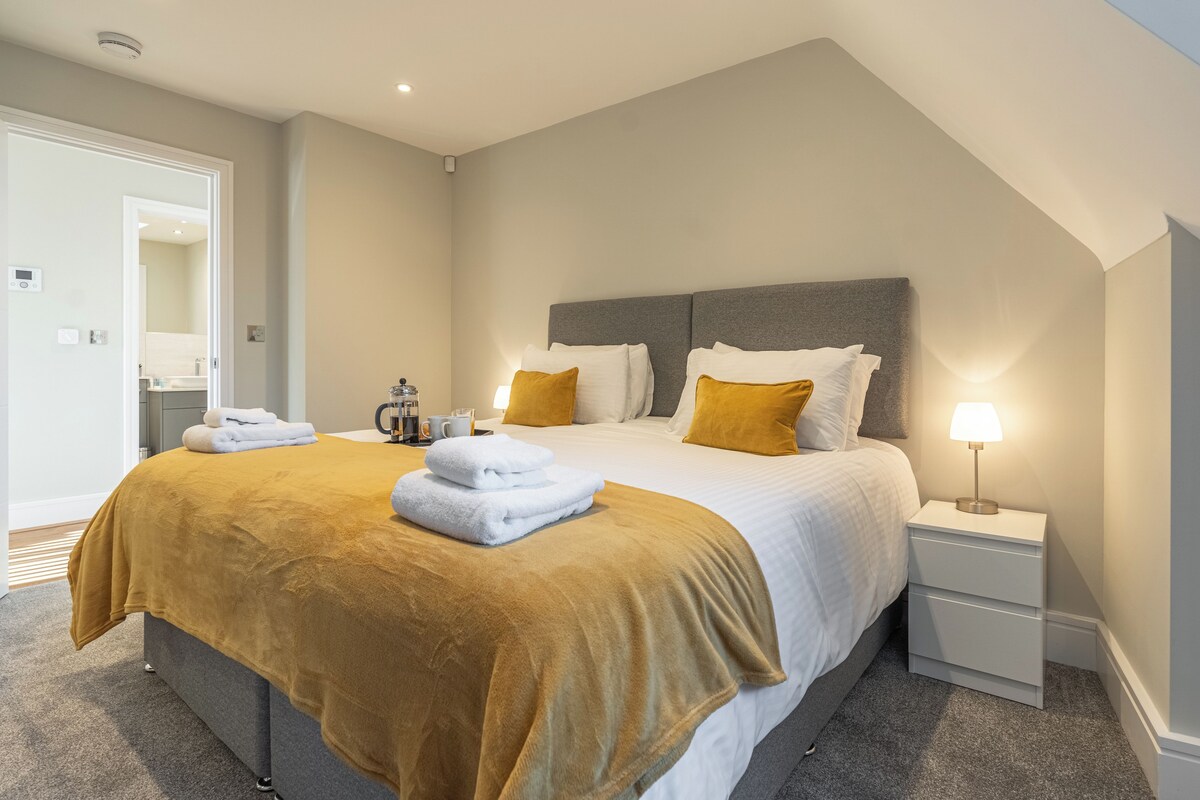 A neatly made bed with a brown blanket, white pillows, and a bedside lamp. There is a small bedside table with a lamp and a cup. The room has a simple, clean design with a gray headboard and a door leading to another room.