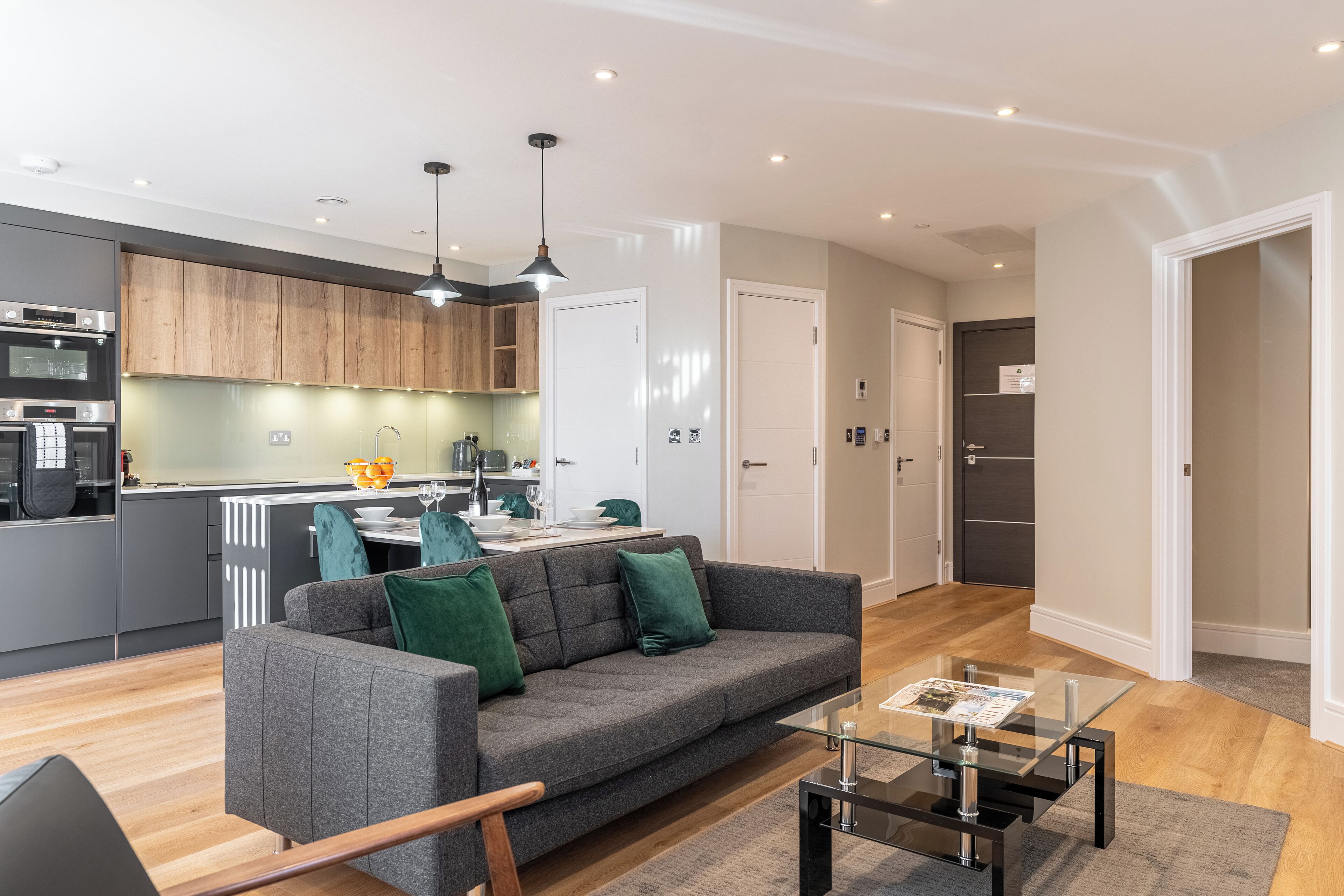 A modern kitchen with wooden cabinets, a dining area with a table and chairs, a grey sofa with green cushions, and a glass coffee table.