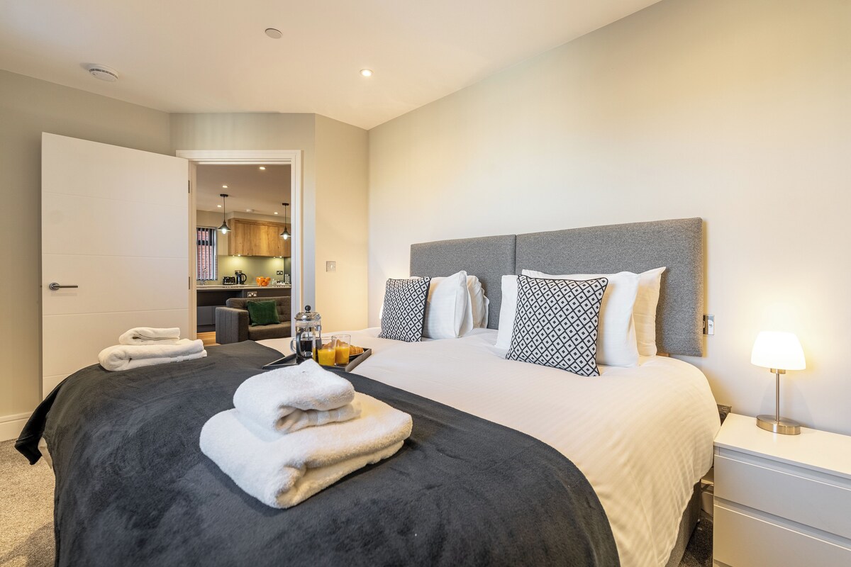 Quarto de hotel com uma cama grande, mesa de cabeceira com abajur e vista para a cozinha.