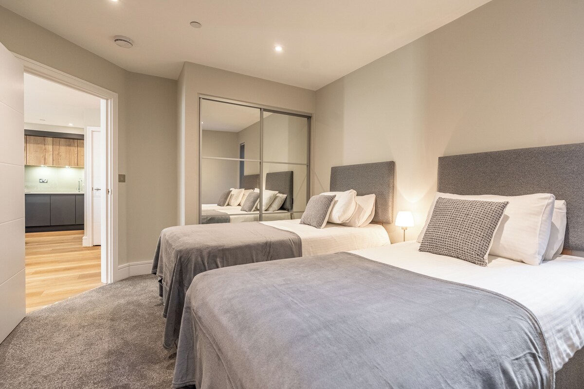 A modern bedroom with two beds, a large mirror, and a view of a kitchen.
