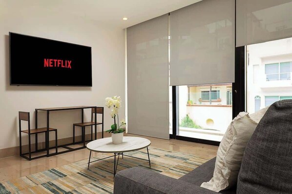 A modern living room with a flat-screen TV displaying the Netflix logo, a round coffee table with a vase of flowers, a gray sofa, and a view of a building through a large window.