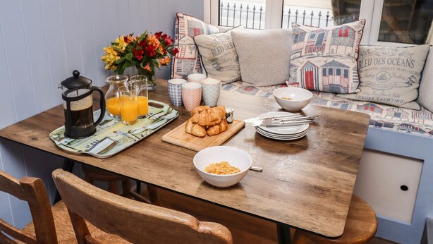 Une table en bois dressée pour le petit-déjeuner, avec une cafetière, des verres de jus d’orange, des croissants et un bol de céréales.