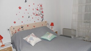 A neatly made bed with two pillows, a wall decal of a tree and flowers, a red lamp on a bedside table, and a white curtain partially drawn to the side.