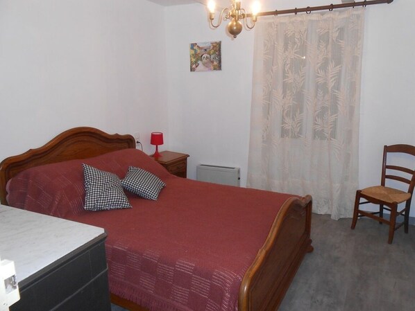 A bedroom with a wooden bed, a red blanket, a checkered pillow, a red lamp, a chair, and a radiator.