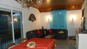 A dining area with a red tablecloth, wooden chairs, a sofa with cushions, a small table, and a kitchen area with a cabinet and counter.
