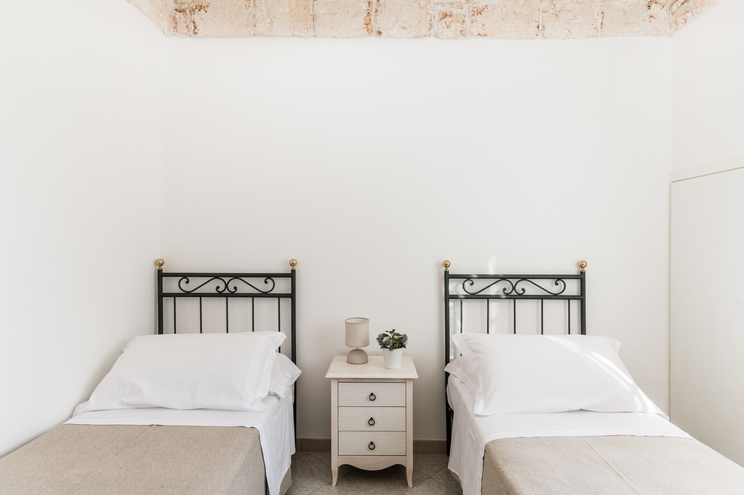 Una habitación con dos camas individuales, una mesita de noche pequeña con una lámpara y una planta, y una pared blanca lisa.