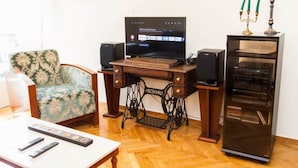 A living room with a wooden desk, a television, a vintage sewing machine, a speaker, a decorative candelabra, and a floral-patterned armchair. - Renovated apt in the Heart of Piraeus (Piraeus)