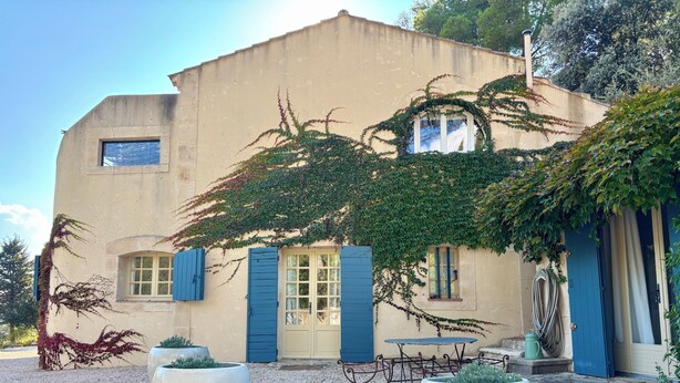 Un bâtiment à la façade beige, aux volets bleus et doté d’une petite fenêtre. On peut y voir deux grands jardinières devant l’entrée.