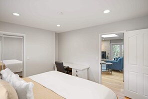 A bedroom with a bed, a desk, and a chair. There is a door leading to a living area with a sofa and a table.