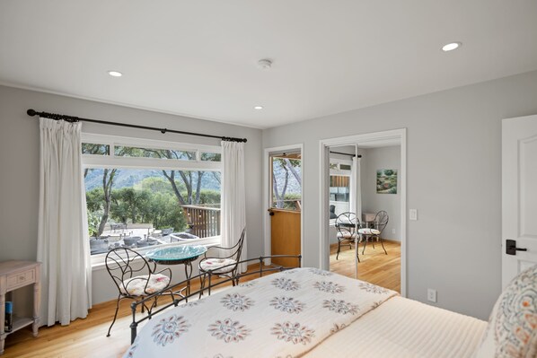 A bedroom with a bed, a window with curtains, a balcony with outdoor furniture, and a door leading to another room.