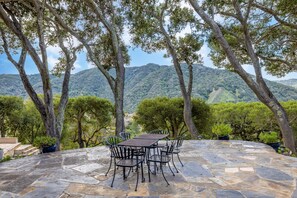 Eine Terrasse im Freien mit Steinböden, Metallstühlen und einem Tisch, umgeben von Bäumen und einer Bergkulisse.