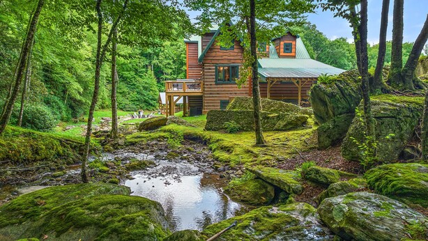 Een rustieke houten hut, omgeven door weelderige begroeiing en een kleine beek.