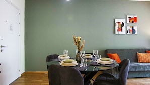 Ein Essbereich mit einem Glastisch, gedeckt für vier Personen, umgeben von dunklen Stühlen. Gegen die Wand lehnt ein graues Sofa mit orangefarbenen Kissen, darüber hängt ein abstraktes Kunstwerk.