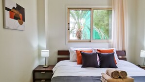 Ein ordentlich bezogenes Bett mit dunkler Tagesdecke, zwei Kissen und einem aufgerollten Handtuch. Ein Fenster mit Vorhängen, ein Nachttisch mit Lampe und eine Klimaanlage an der Wand sind ebenfalls zu sehen.