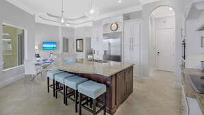 A modern kitchen with a central island, bar stools, a large refrigerator, and a dining area with a TV. - SW Exposure Home | Wide Canal w/ Pool & Spa (Marco Island)