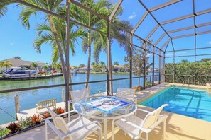 A covered outdoor pool area with a glass table and chairs, overlooking a body of water and palm trees.
