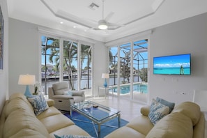 A spacious living room with a large window overlooking a pool and palm trees, a ceiling fan, and a flat-screen TV mounted on the wall.