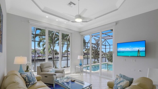 A spacious living room with a large window overlooking a pool and palm trees, a ceiling fan, and a flat-screen TV mounted on the wall. - SW Exposure Home | Wide Canal w/ Pool & Spa (Marco Island)