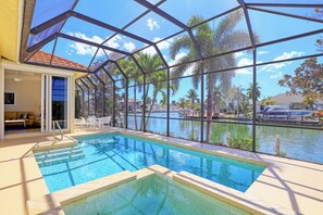 Zona de piscina con cerramiento de vidrio, una silla de descanso y vistas a una masa de agua con palmeras.
