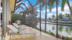 A sunlit outdoor lounge area with white lounge chairs and a grill, overlooking a body of water and palm trees. - SW Exposure Home | Wide Canal w/ Pool & Spa (Marco Island)