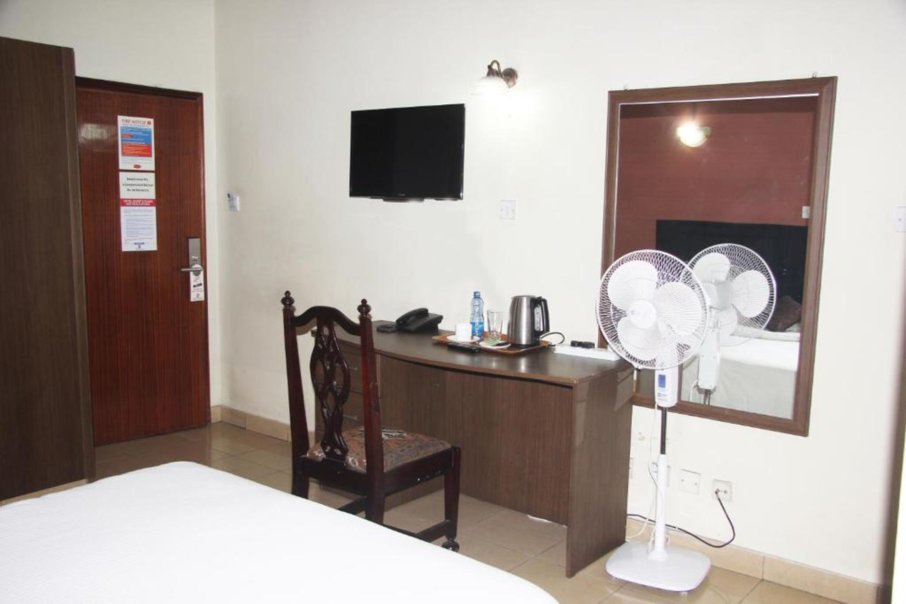 A hotel room with a bed, a desk with a chair, a television, a wall-mounted fan, and a door with a sign.