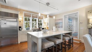 A modern kitchen with a central island, bar stools, a refrigerator, and a view of the outdoors through large windows.