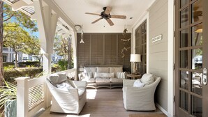 A covered porch with white furniture, a ceiling fan, and a sign reading "BEACH HOUSE".