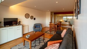 A modern living room with a sofa, coffee table, and a TV on a cabinet. There is a dining area with a table and chairs. - Quinta do Mar - Watch The Sun Rise Over The Ocean (Angra do Heroismo)