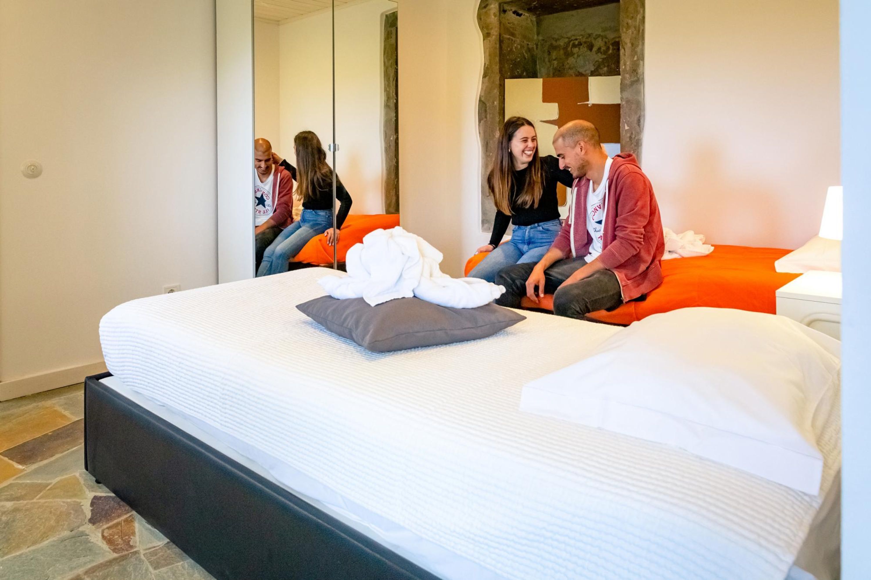 A hotel room with a large bed, orange accent pillows, a white comforter, and a stone-patterned floor.