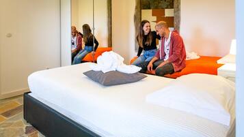 A hotel room with a large bed, orange accent pillows, a white comforter, and a stone-patterned floor.