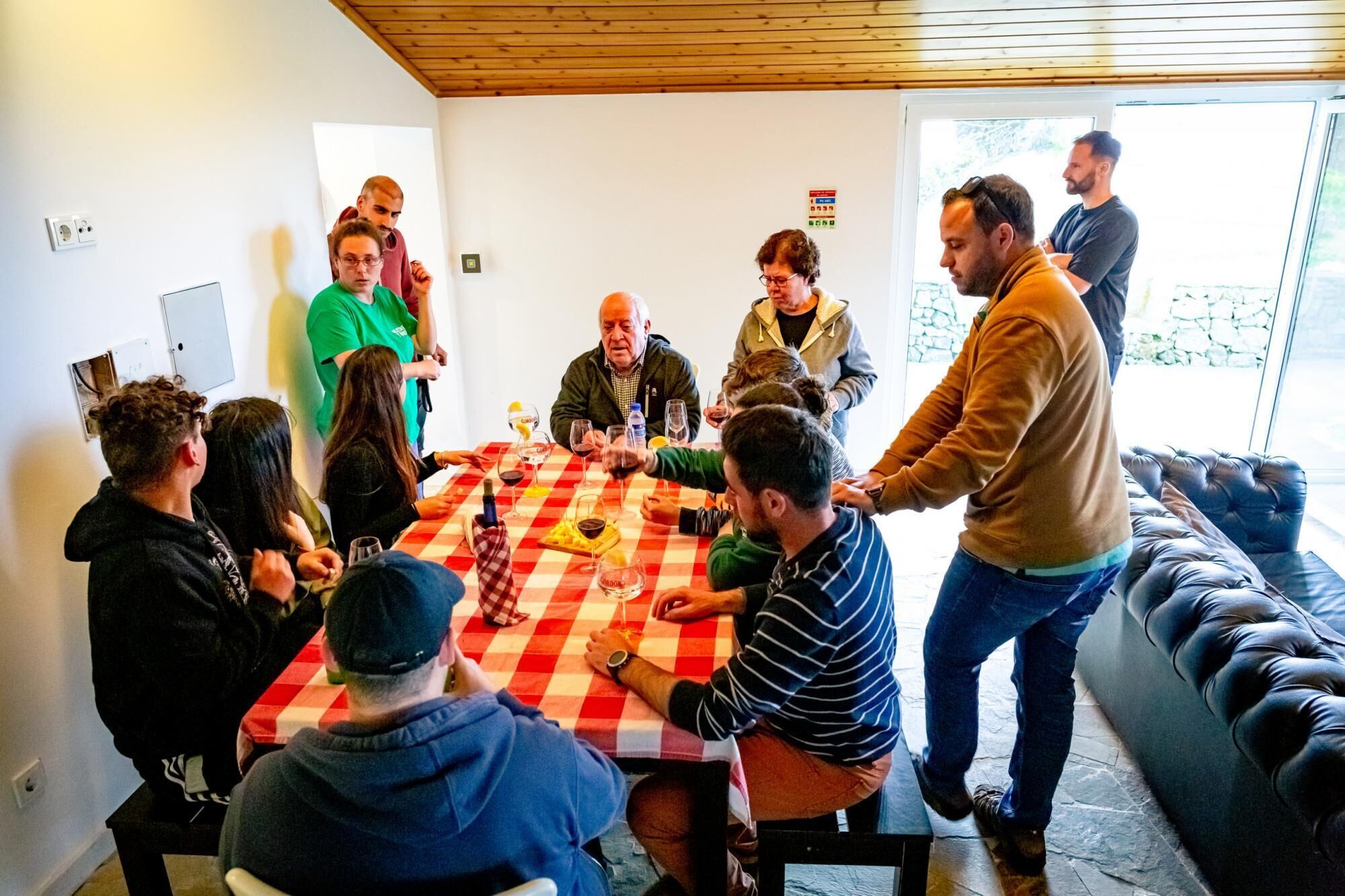 Un grup de persones assegudes al voltant d'una taula amb un mantell de taula vermell i blanc a escacs, conversant.