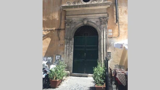 A building with a large arched entrance, a small circular window above the door, and a plaque with numbers. There are potted plants and a table with chairs outside. - Piazza Navona Penthouse - Piazza Navona Penthouse (Rome)
