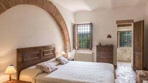 A bedroom with a large bed, a wooden headboard, a nightstand with a lamp, a dresser, and a window with a view of greenery.