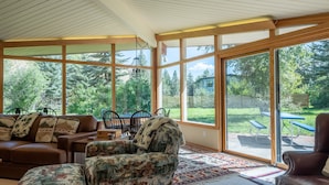 Un salón soleado con ventanas amplias, un sofá de cuero marrón, un sillón estampado y un comedor con mesa y sillas.