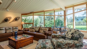 Una sala de estar acogedora con un sofá de cuero marrón, un sillón estampado con motivos florales y una mesa de centro de vidrio. Amplias ventanas ofrecen vistas a un jardín exuberante.