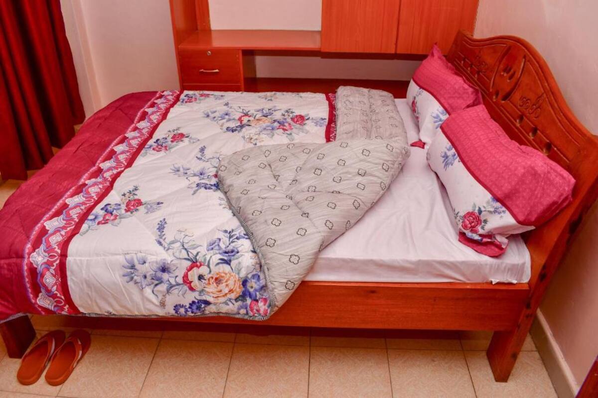 Ein hölzernes Bett mit floralem Überwurf und Kissen, ein Nachttisch aus Holz und ein Paar Slipper auf dem Boden.