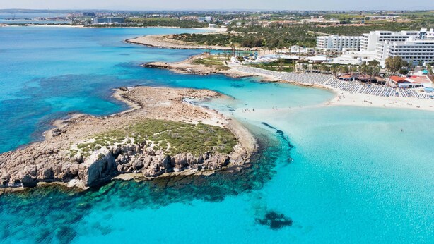 An aerial view of a coastal area with clear turquoise waters, rocky outcrops, and a beach with sun loungers. - Protaras holiday villa - rentprivatevillas 113 (Protaras)