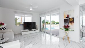 A modern living room with a flat-screen TV, a ceiling fan, and a large sliding glass door leading to a pool area. - Protaras holiday villa - rentprivatevillas 113 (Protaras)