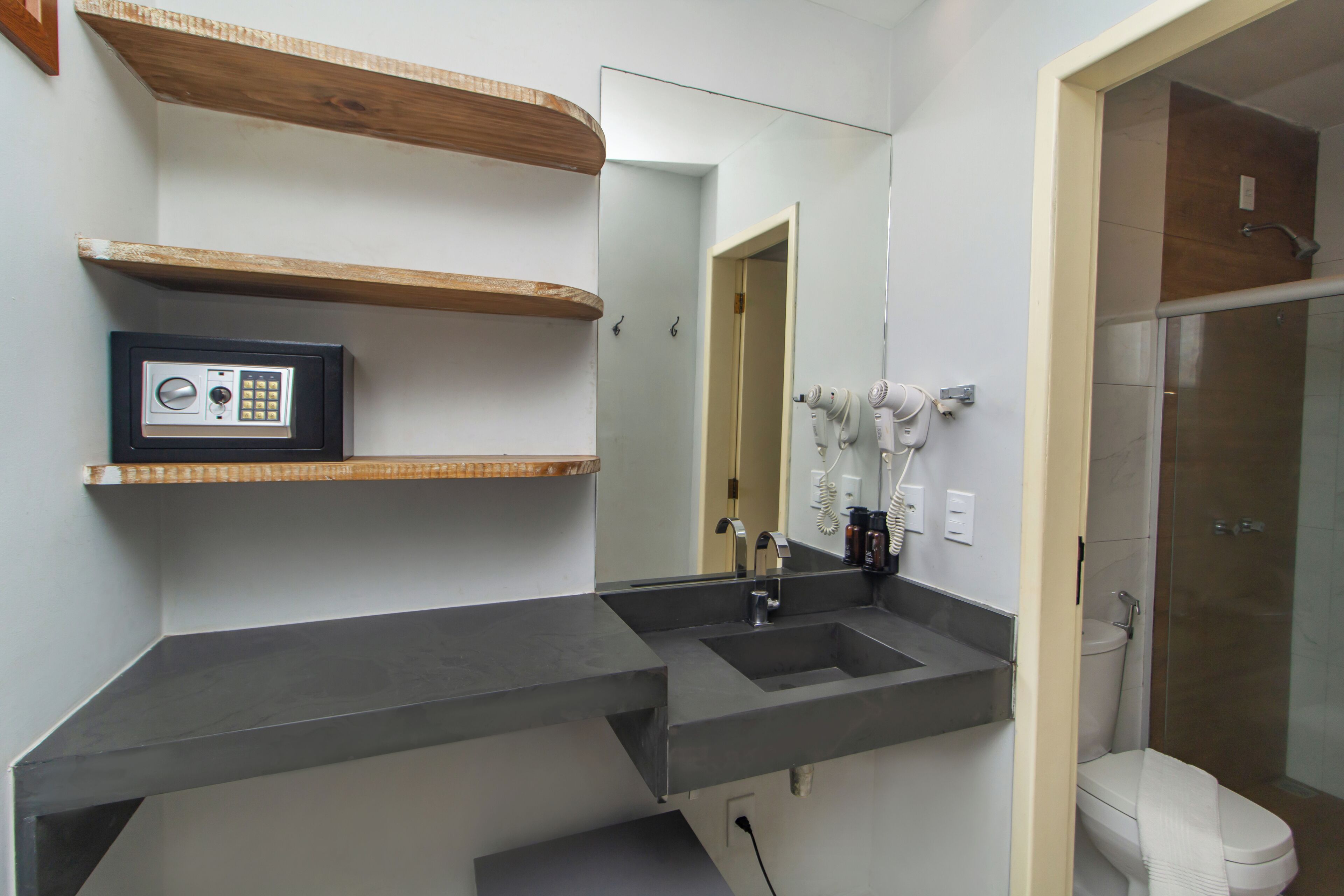 Une salle de bain avec un grand miroir, un lavabo, des toilettes et une douche. Il y a des étagères en bois au-dessus du lavabo et un coffre-fort sur la gauche.