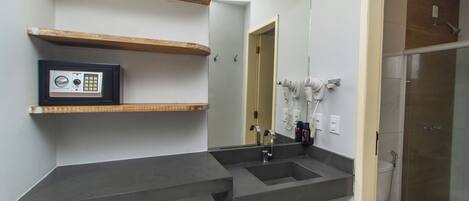 Une salle de bain avec un grand miroir, un lavabo, des toilettes et une douche. Il y a des étagères en bois au-dessus du lavabo et un coffre-fort sur la gauche.