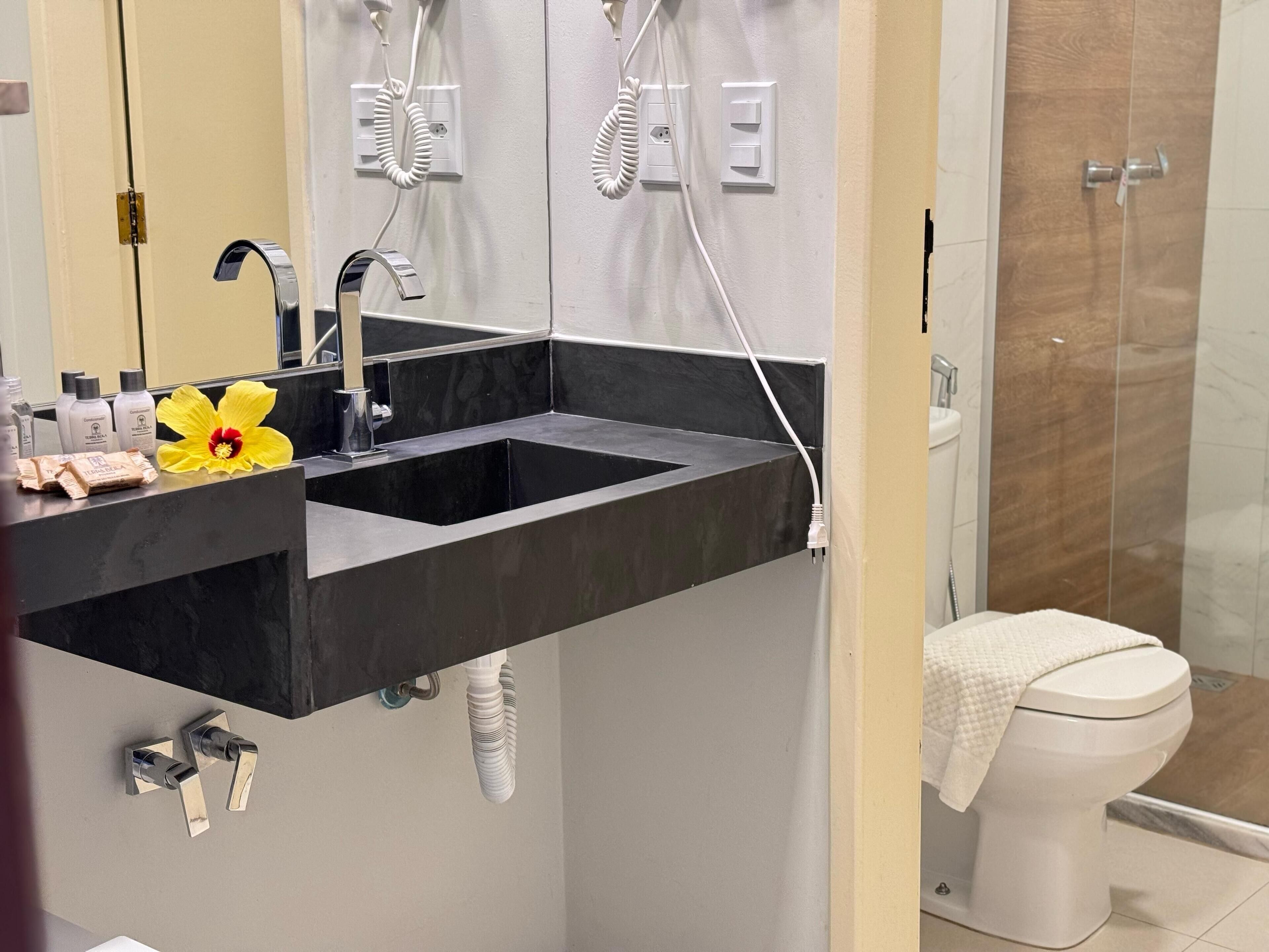 Een badkamer met een zwarte wastafel, een spiegel en een toilet. Er staan toiletartikelen op de wastafel en een bloem op het aanrecht.