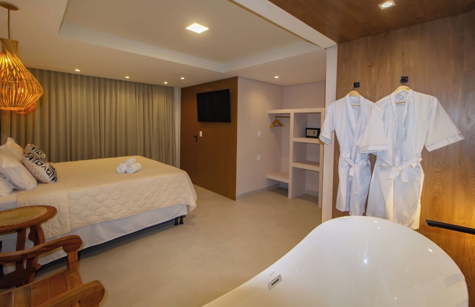 Een hotelkamer met een bed, een houten stoel, een kledingrek met badjassen, een televisie en een bad.