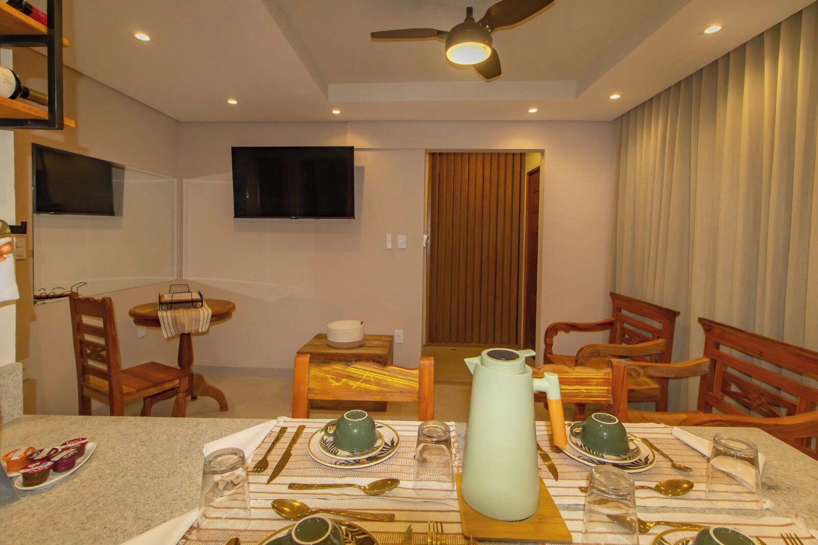 A dining area with a table set for four, a television mounted on the wall, and a ceiling fan.