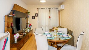 A dining area with a round table set for four, surrounded by white upholstered chairs. A television is mounted on the wall above the table, and there are decorative items on shelves and a small table. - LOCAR-IN GRAMADO- Vista do Quilombo (Gramado)