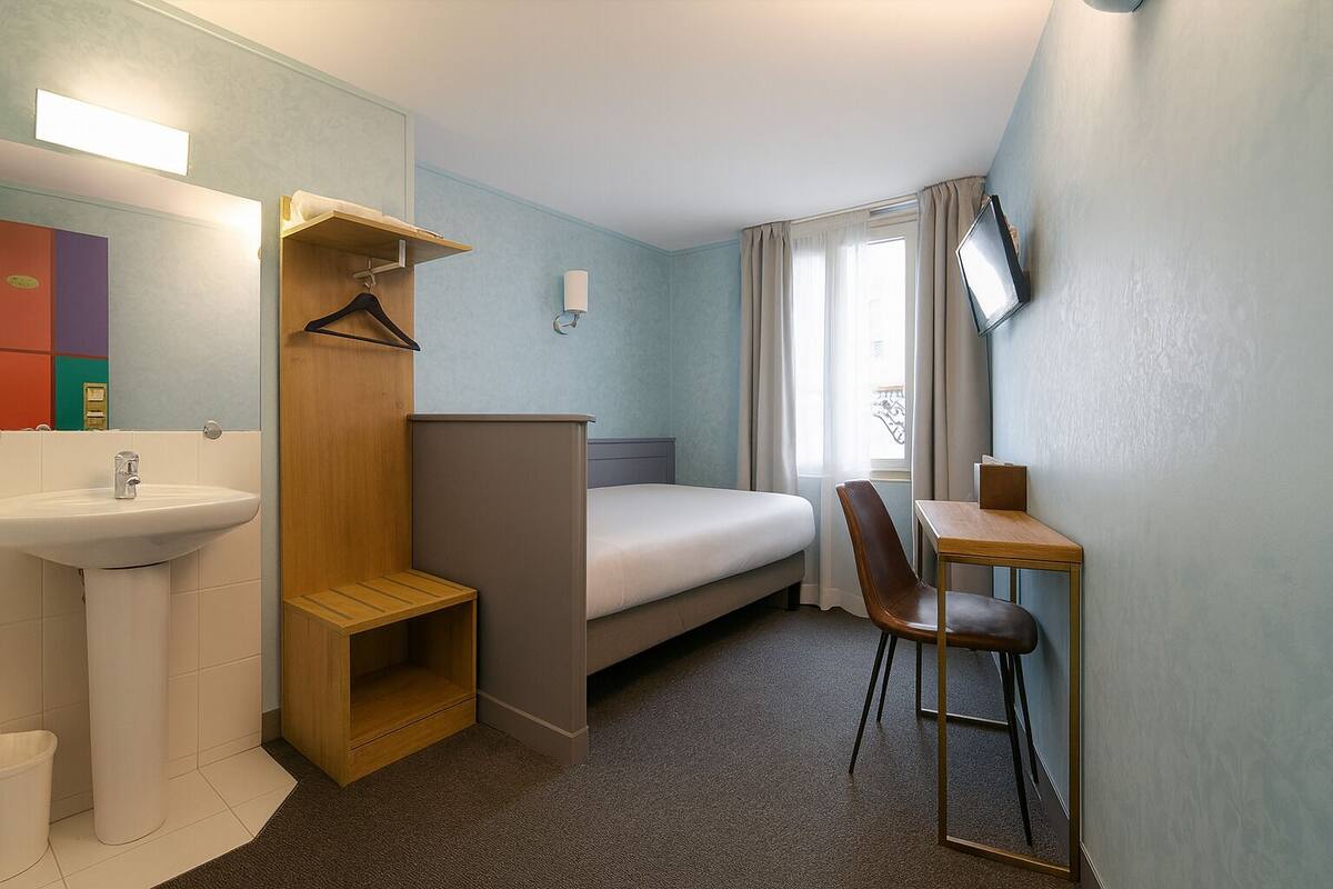 A compact hotel room with a single bed, a small desk with a chair, a bedside table, a mirror, a sink, and a wardrobe.