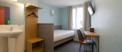Una habitación de hotel compacta con una cama individual, un escritorio pequeño con silla, una mesita de noche, un espejo, un lavamanos y un armario.