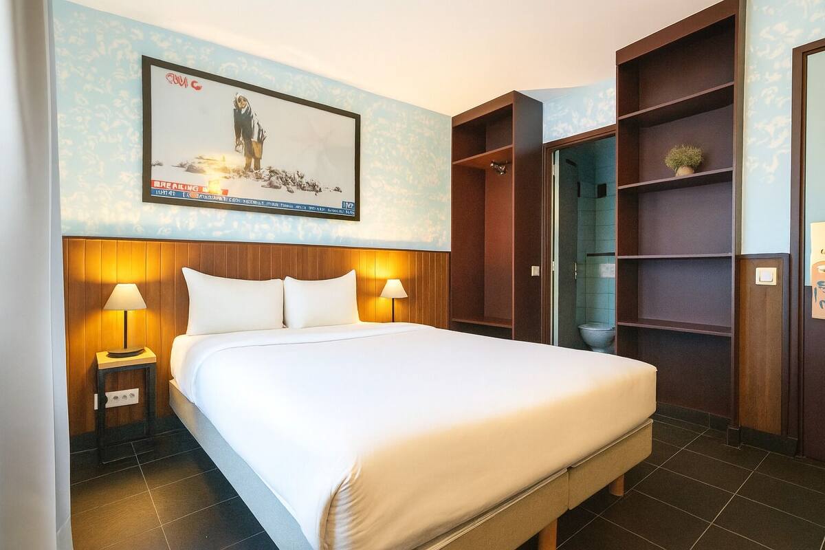 A hotel room with a large bed, wooden headboard, two bedside lamps, a poster on the wall, and a built-in wardrobe with shelves.