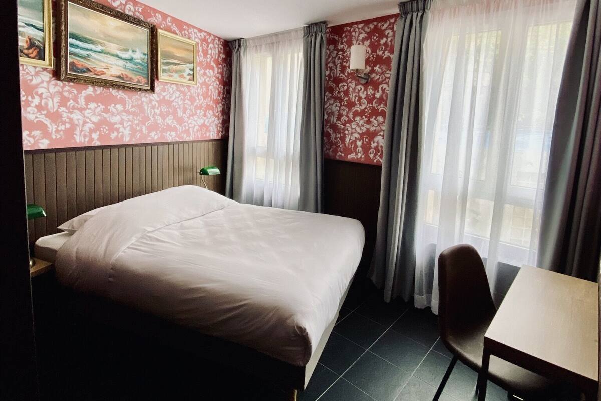A hotel room with a bed, a desk, a chair, a lamp, and a painting on the wall.